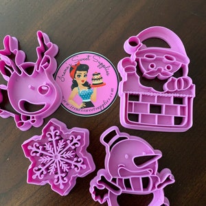 May include: Set of four purple plastic cookie cutters, featuring a reindeer, Santa Claus in a chimney, a snowflake, and a snowman. The cutters are on a brown wooden surface.