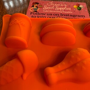 Puede incluir: Molde de silicona naranja con seis cavidades para hacer varios alimentos. Las cavidades tienen forma de lata, hamburguesa, pierna de pollo, cono y otras dos formas. El molde está sobre una superficie de madera.