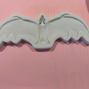 May include: A white bat-shaped mold with outstretched wings. The mold has detailed wing and body textures. The bat is centered and appears to be made of a hard, durable material. The background is a solid pink color.