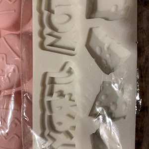 May include: White silicone mold with the words "MOM" and "DAD" in raised lettering. The mold also has three smaller shapes, each with a different design.