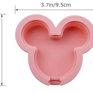 May include: A light pink silicone mould shaped like a mouse head with rounded ears. The mould features a recessed centre and raised edges. Dimension lines indicate a width of 9.5 cm and a height of 8 cm for the overall structure.