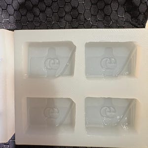 Puede incluir: Un molde de silicona blanco con cuatro cavidades rectangulares, cada una con un logotipo de Etsy grabado. El molde tiene un acabado mate y liso, y está colocado sobre un fondo de tela gris oscuro con una textura hexagonal de panal.