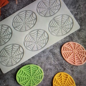 May include: A silicone mold with six oval-shaped spiderweb designs. Three finished spiderweb-shaped decorations in green, peach, and yellow are also present. The items are likely for Halloween-themed crafts or baking.