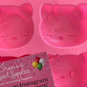 May include: A pink silicone mold featuring three Hello Kitty face designs. The mold is for cake decorating and has a business card with the text "Irene's Sweet Supplies" and social media information.