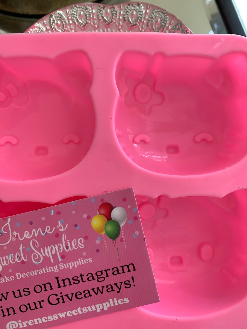 May include: A pink silicone mold featuring three Hello Kitty face designs. The mold is for cake decorating and has a business card with the text "Irene's Sweet Supplies" and social media information.