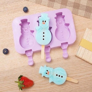 May include: Purple silicone mould with three snowman-shaped popsicle moulds. The mould has a stick holder for each popsicle. There are three blue snowman popsicles, two in the mould and one on a wooden stick.