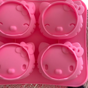 May include: A pink silicone mold featuring four Hello Kitty face designs. Each face has a bow, eyes, nose, and whiskers. The mold is designed for making decorative treats or crafts. The background includes a silver patterned surface and a promotional card.