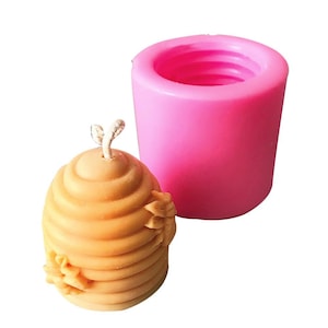 May include: A pink silicone mold for making a beehive-shaped candle. The mold is cylindrical and has a smooth, shiny surface. The candle is made of a light brown wax and has a detailed beehive design with small bees on the sides.