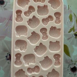 May include: A light pink silicone ice cube tray with various shaped molds, including bows and cat faces. The tray is rectangular and features multiple cavities for creating themed ice cubes. The design is suitable for making fun-shaped treats.