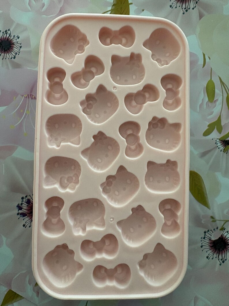 May include: A light pink silicone ice cube tray with various shaped molds, including bows and cat faces. The tray is rectangular and features multiple cavities for creating themed ice cubes. The design is suitable for making fun-shaped treats.