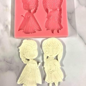 May include: A pink silicone mold with two cartoon princess designs, and two finished cream-colored princess figures. The mold features detailed princess figures with flowing dresses and braided hair. The finished figures are standing.