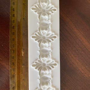 May include: A white silicone mold featuring a repeating ornate floral design. The mold is approximately 10 inches long and includes a ruler for scale. The design is symmetrical and detailed, suitable for crafting projects.