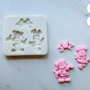 以下が含まれることがあります： サンタの帽子をかぶったミッキーマウスのキャラクター2人と「JOY」の文字のデザインが施された、3つのクリスマスをテーマにしたデザインのシリコンモールド。モールドの隣には、サンタの帽子をかぶったミッキーマウスのキャラクター2人と「JOY」の文字のデザインが施された、ピンク色の完成したレジン作品3つがあります。