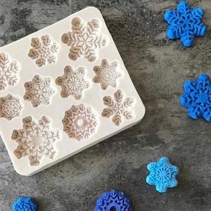 Puede incluir: Un molde de silicona blanco con 10 diseños de copos de nieve. El molde está sobre una superficie gris texturizada. Hay 6 objetos azules en forma de copo de nieve al lado del molde.