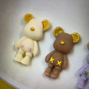 May include: Close-up of three small bear figurines. The bears are in cream, brown, and purple. Each bear has gold-colored ears and yellow or green accents. The cream bear has pink accents, and the brown bear has yellow accents.