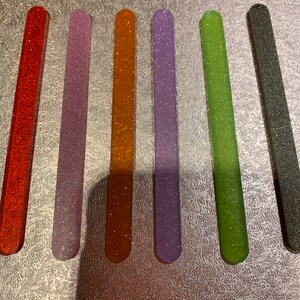 May include: Six nail files with glitter embedded in the surface. The files are in the colours red, purple, orange, lavender, green, and grey.