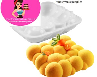 May include: A white silicone mould for baking a cake with a bubble design. The mould is shown with a yellow cake baked in it. The cake is decorated with a sprig of mint and a few orange slices.