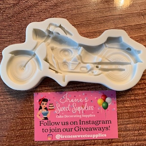 May include: White silicone mould of a motorcycle. The mould is designed to create a cake topper or decoration. The mould is on a wooden surface.