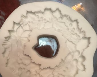 Puede incluir: Un molde de silicona blanco de forma circular que presenta un patrón detallado en relieve de hojas y bayas de acebo dispuestas en forma de corona. El centro del molde tiene un recorte circular liso y el diseño general es texturizado.