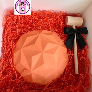 Op de afbeelding: Een grote oranje chocoladesfeer met een geometrisch ontwerp, een houten hamer en een zwarte strik, allemaal in een doos gevuld met oranje geribbeld papier.