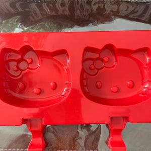 May include: A red silicone mold for making popsicles, featuring two Hello Kitty face designs. The mold has a rectangular shape with two slots for the popsicle sticks. The design includes the iconic bow and facial features.
