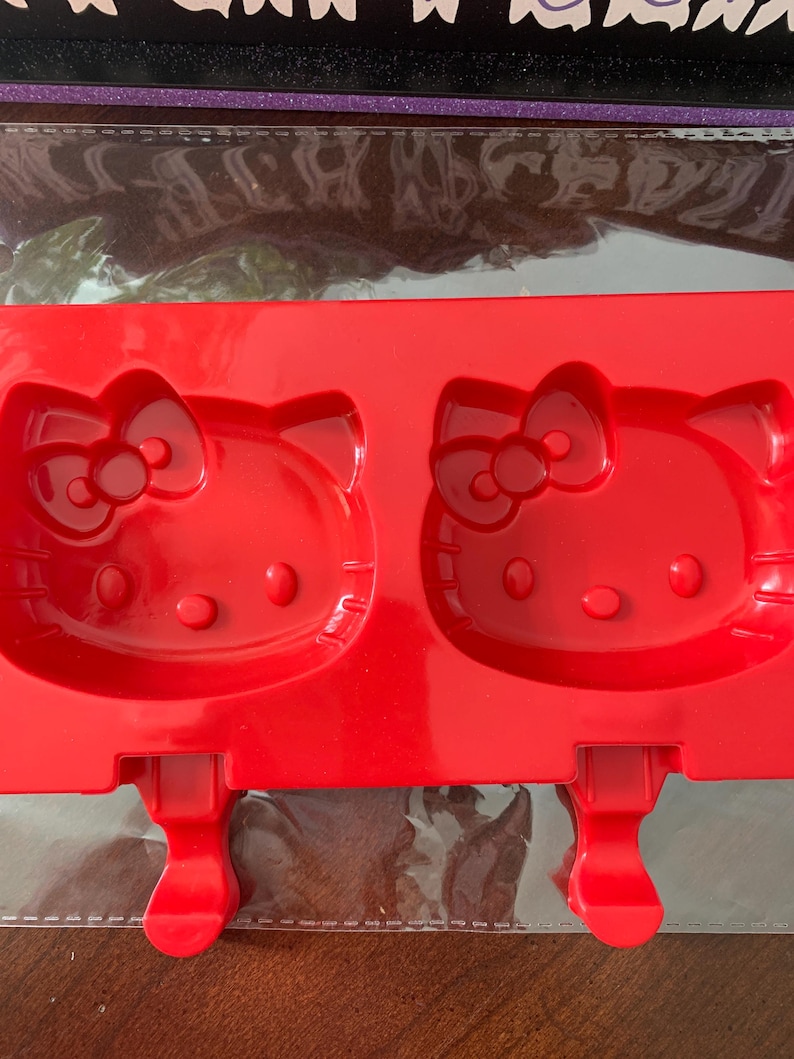 May include: A red silicone mold for making popsicles, featuring two Hello Kitty face designs. The mold has a rectangular shape with two slots for the popsicle sticks. The design includes the iconic bow and facial features.