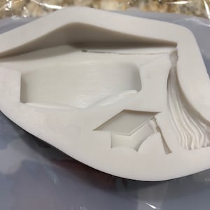 May include: White silicone mold with a graduation cap design. The mold is designed to create a cake topper or other decorative item.