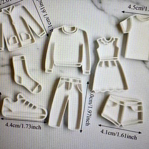 以下が含まれることがあります： ジャケット、セーター、ドレス、Tシャツ、靴下、スニーカー、パンツ、ショートパンツの形をした白いプラスチック製のクッキー型セット。ジャケットの5.1cmやスニーカーの4.4cmなど、センチメートル単位で寸法が明記されています。