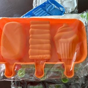 May include: Orange silicone popsicle molds in various shapes, including a classic popsicle, a layered popsicle, and a cupcake. Additional molds in blue and white are visible in the background. The molds are packaged in clear plastic.