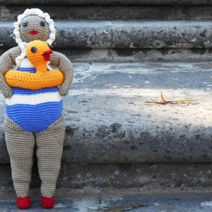 Puede incluir: Muñeca de ganchillo hecha a mano con un traje de baño a rayas azules y blancas y un flotador de pato amarillo y naranja. La muñeca tiene un gorro blanco y zapatos rojos. La muñeca está sobre unos escalones de piedra.