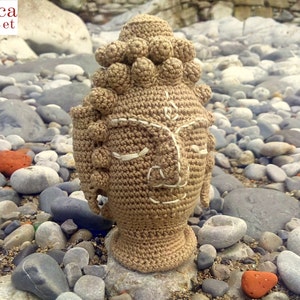 Puede incluir: Cabeza de Buda de ganchillo marrón con puntadas blancas para los ojos y la boca. La cabeza está sentada sobre un lecho de pequeñas piedras grises.