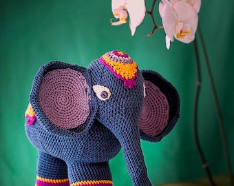 Op de afbeelding: Een gehaakt blauw olifantenspeelgoed met paarse oren, gele en roze accenten en een wit oog. De olifant staat op een lichtblauw oppervlak met een reflectie van de olifant zichtbaar.