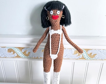 May include: A crocheted doll with brown skin, black hair, and white clothing. The doll is wearing a white one-piece swimsuit and white boots.