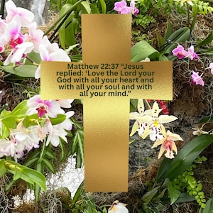 May include: A gold cross with textured surface, surrounded by vibrant orchids in shades of white, pink, and yellow. The text on the cross reads: Matthew 22:37 "Love the Lord your God with all your heart and with all your soul and with all your mind."