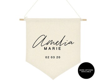 May include: A beige canvas banner with a wooden dowel at the top and a white string for hanging. The banner has black text that reads "Amelia Marie 02 03 20".