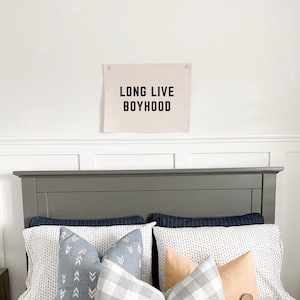 Op de afbeelding: Een grijs hoofdbord met een donkerblauw en wit beddengoed. Er zijn verschillende kussens op het bed, waaronder een grijs en wit geruit kussen, een lichtblauw kussen met een chevron-patroon en een perzikkleurig kussen met een knoop. Een wit canvas-banner met de tekst "LONG LIVE BOYHOOD" hangt aan de muur boven het bed.