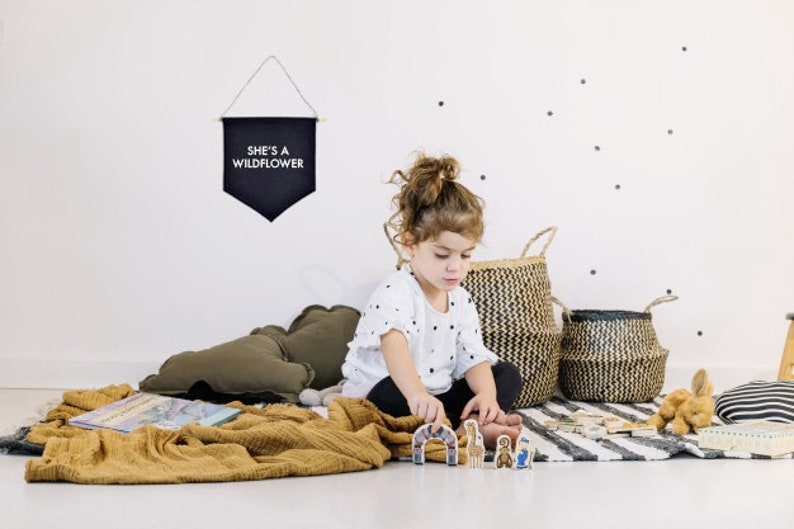May include: A young girl plays with wooden toy figures on a rug with black and white stripes. The rug is surrounded by pillows and baskets. A black banner with white text that reads "She's a Wildflower" hangs on the wall behind the girl.