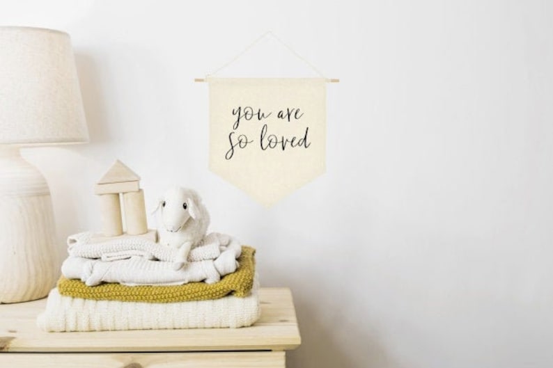 May include: A white canvas banner with the text "you are so loved" hanging on a white wall. The banner is decorated with a simple wooden dowel at the top and bottom.