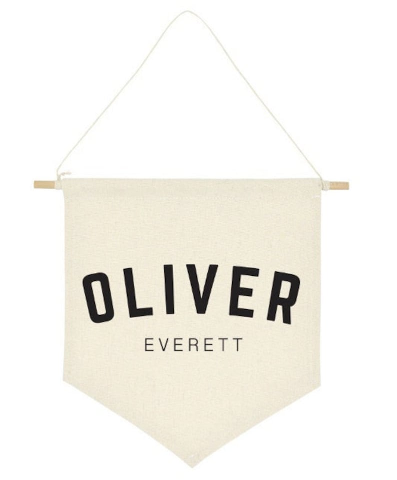 May include: A white canvas banner with the name "OLIVER EVERETT" printed in black. The banner is hanging from a wooden dowel rod with a white string.