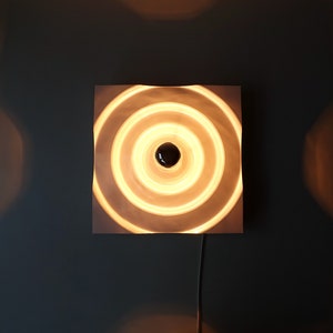 Puede incluir: Una l&aacute;mpara de pared blanca con un dise&ntilde;o circular. La l&aacute;mpara tiene c&iacute;rculos conc&eacute;ntricos que crean un efecto de ondulaci&oacute;n. La luz est&aacute; encendida y emite un brillo blanco c&aacute;lido.