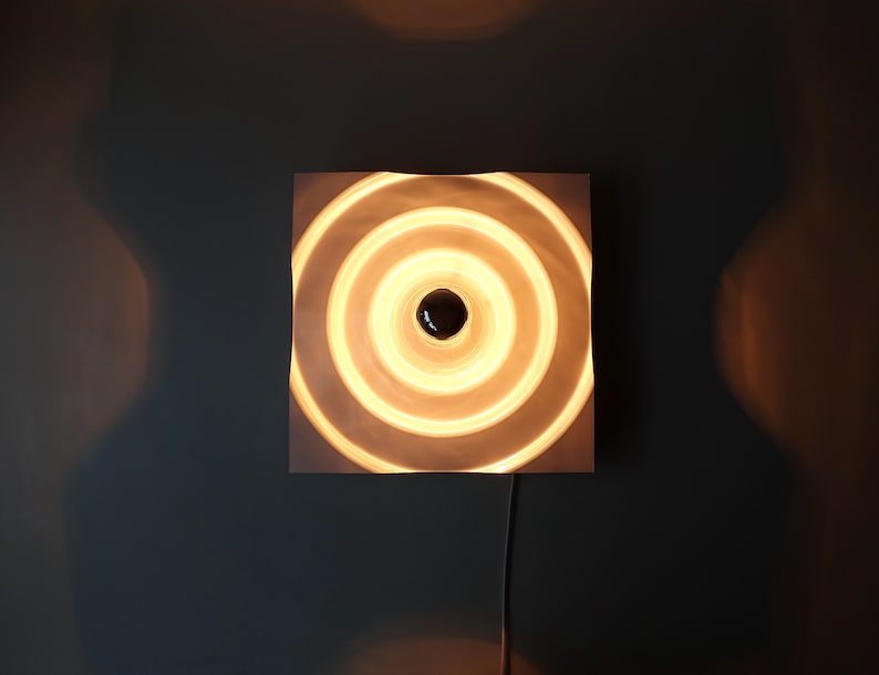 Puede incluir: Una l&aacute;mpara de pared blanca con un dise&ntilde;o circular. La l&aacute;mpara tiene c&iacute;rculos conc&eacute;ntricos que crean un efecto de ondulaci&oacute;n. La luz est&aacute; encendida y emite un brillo blanco c&aacute;lido.