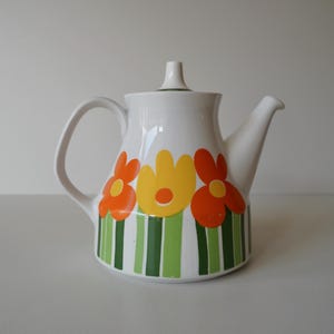 Può includere: Teiera in ceramica bianca con un design floreale. La teiera ha un corpo arrotondato con un beccuccio e un manico. I fiori sono arancioni, gialli e verdi.