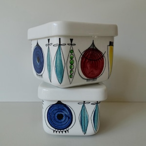 May include: Two white ceramic square storage containers with lids. The containers have a geometric design with blue, green, red, and yellow accents. The design features circles, leaves, and other abstract shapes.