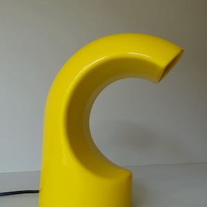 Op de afbeelding: Een felgele tafellamp met een gebogen, abstract ontwerp. De lamp heeft een gladde, glanzende afwerking.