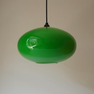 Könnte beinhalten: Eine leuchtend grüne, ovale Pendelleuchte. Die glänzende Oberfläche reflektiert die Umgebung. Die Leuchte hängt an einem schwarzen Kabel vor einem neutralen Hintergrund. Diese moderne Leuchte ist ein auffälliges Designelement.