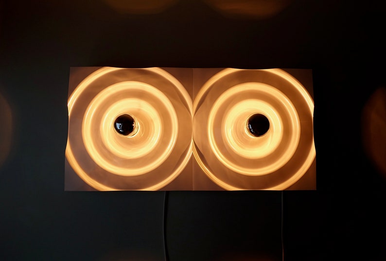 Puede incluir: Una l&aacute;mpara de pared blanca con dos anillos circulares conc&eacute;ntricos. Los anillos est&aacute;n iluminados con luz blanca c&aacute;lida.