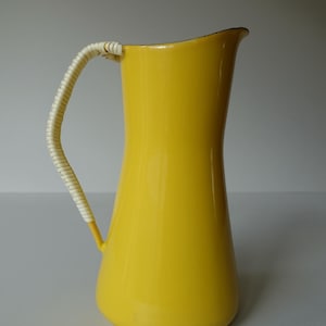 Puede incluir: Una jarra de esmalte amarillo con un asa tejida blanca. La jarra tiene una base ancha y un cuello estrecho.