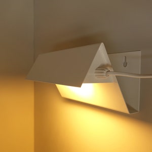 May include: A white triangular wall lamp with a cord and plug. The lamp is switched on and emits a warm yellow light.