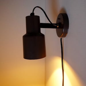 May include: A dark brown wall-mounted lamp with a cylindrical shade and a black cord. The lamp is attached to a circular base plate and emits a warm, downward-facing light that illuminates the wall below the fixture in a soft golden glow.