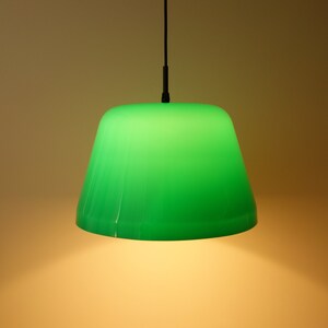 Könnte beinhalten: Eine grüne Pendelleuchte mit einem konischen Lampenschirm. Der Schirm ist leuchtend grün und hängt an einem schwarzen Kabel. Das Licht ist eingeschaltet und erzeugt einen warmen Schein darunter.